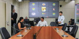 Dois parlamentares sentados à mesa, em reunião presencial, acompanhados por parlamentares que participam remotamente, exibidos em telão, ao fundo.