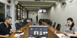 dois parlamentares sentados à mesa, em reunião presencial, e dois participando remotamente, exibidos em tela de computador.ticipando 