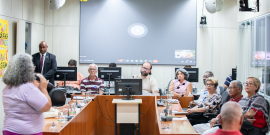 Quatro homens e três mulheres reunidos, sentados à mesa, assistem a mulher que fala, de pé.