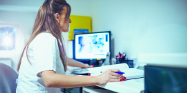 Sentada e ao lado de computador, mulher de jaleco consulta livro.