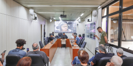 Quatro homens e uma mulher reunidos, sentados à mesa; em telão do fundo, mulher participa de reunião de forma remota. À frente, mais de oito pessoas acompanham a reunião, sentados. 