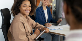 Mulher negra sentada em uma cadeira faz anotações em caderno. Atrás dela está uma mulher mais velha de cabelos grisalhos longos, também fazendo anotações. À frente dela, de costas e desfocada, está uma pessoa em pé, ensinando.