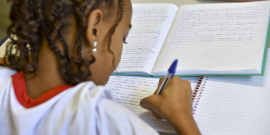 Criança negra de uniforme, de costas sentada na carteira, escreve em caderno com caneta azul.