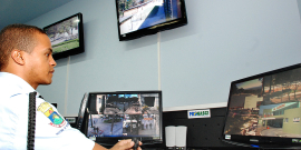 Telas mostram imagens de videomonitoramento da cidade; guarda municipal senta em frente ao computador