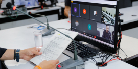 Tela do computador mostra reunião virtual dos vereadores. Em frente à tela, duas mãos seguram um documento com a pauta da reunião.