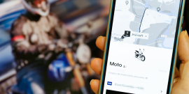 Em primeiro, plano, celular com aplicativo de corrida por moto. Em segundo plano, motociclista