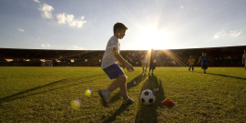 Menino chuta bola em campo de futebol próximo