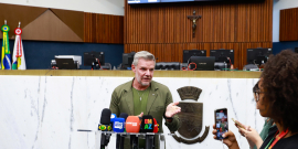 Foto do vereador Professor Juliano Lopes, que aparece centralizado falando com repórteres  