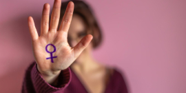 Mulher estende a palma da mão na frente de seu rosto. A palma de sua mão tem o símbolo do sexo feminino pintada de roxo