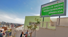 Fachada do Centro de Saúde Nossa Senhora Aparecida com placa indicativa. Rua em declive. Dua mulheres na porta