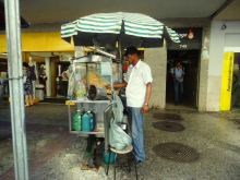 Pipoqueiro está parado junto a seu carrinho de pipocas, em frente a estabelecimento comercial