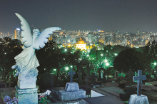 Vista noturna de lápides e esculturas no Cemitério do Bonfim. Ao fundo, luzes da cidade. Vista panorâmica da cidade