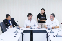 Integrantes da Comissão estão sentados à Mesa apreciando a pauta da reunião. Atrás deles, de pé, uma servidora assessora o presidente.