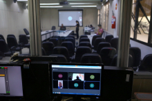 Tela de computador exibindo videoconferencia. Ao fundo, cadeiras vazias no pplnaéio e telão exibindo videoconferência
