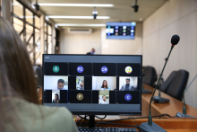 Monitor de computador exibe vereadores em videoconferência. Ao fundo, cadeiras vazias do Plenário Camil Caram