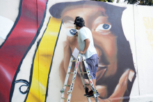 Artista no alto da escada grafitando muro. Muro parcialmente pintado, exibe rosto e faixas coloridas