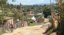 Estrada de terra com material de construção à direita e homem de bicicleta à esquerda. Nos dois lados, barrações com tijolos aparentes, durante o dia. 