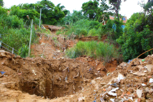 Barranco com terra e vegetação nas laterais; no alto, ao fundo, uma residência. Imagem diurna. 