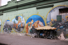 Em frente a um muro pintado com imagens de homens trabalhando, há trêsx carrinhos de supermercado cobertos com um colchão, indicando que há um pessoa habitando o local.