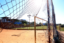 Trava do gol em um campo de futebol, com a rede rasgada. O campo é de terra vermelha e o alambrado está danificado severamente