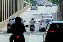 Duas motos e três carros trafegam em viaduto, durante o dia. Uma das motos está entre dois carros. 
