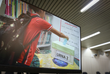 imagem na tela do computador mostra criança ,vestida com camisa vermelha e usando mochila preta nas costas, votando em uma urna  de papelão. Ao fundo, uma estante cheia de livros e um quadro branco nos remete á ideia de sala de aula 