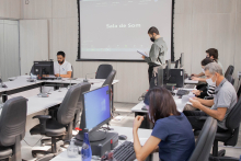 Vereador sentado à mesa, preside reunião semipresencial. Em plenário, cinco servidores da Câmara auxiliam na reunião