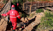 Dois funcionários de obras com uniformes e capacetes vermelhos andar por obra de grande porte da Prefeitura.