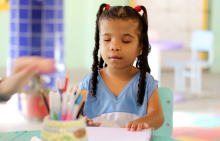 Aluna com deficiência visualpassa suas mãos em caderno em espiral, sentada em carteira escolar. À sua frente, desfocado, um porta lápis com lápis coloridos e mãos femininas. 