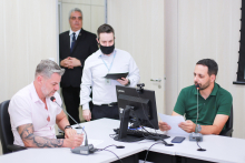 Dois vereadores em reunião presencial sentados à mesa, um deles em frente ao computador. Ao fundo, de pé, dois assessores da Câmara.