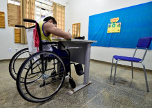 Criança em cadeira de Rodas escreve em carteira escolar, em sala de aula, em frente a um quadro azul.