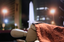 imagem de um boneco de madeira deitado no chão e com um cobertor. Ao fundo, a imagem desfocada do pirulito da Praça Sete