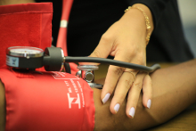 A imagem mostra apenas a mão de uma mulher que mede a pressão arterial com um aparelho vermelho no braço de uma pessoa.
