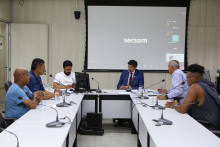 Seis homens sentados à mesa em reunião presencial