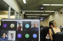 Quatro parlamentares dividem a tela de computador em reunião remota. Ao fundo, Plenário Camil Caram, com assessora da Câmara sentada, acompanhando. 