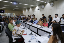 Sala de reunião com doze pessoas sentadas à mesa e mais de vinte sentadas ao fundo e de pé.