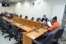 Dois parlamentares sentados à mesa, em reunião, com três homens assistindo , ao fundo.