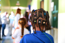 Criança, de costas, com várias tranças no cabelo presas por elásticos de diversas cores. Ao fundo, outra criança e adultos desfocados. 