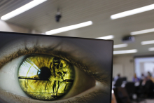 imagem de um olho verde na tela do computador. é possível ver o reflexo de uma mulher sentada de cabeiça baixa diante do punho cerrado de um homem