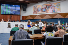 A foto é plano aberto mostra o Plenário Amintas de Barros. Visto de trás, em primeiro plano, está um grupo de vereadores sentados. O vereador Gabriel Azevedo está em pé, virado de frente, falando ao microfone, ele usa blusa social branca e calça jeans. Mais atrás, em um nível acima, estão sentados o presidente da reunião e a secretária, e na parede acima deles o telão que contabiliza a presença e os votos.