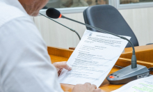 Na foto aparece parte da lateral do braço do vereador, que segura um  papel em que está a pauta da reunião e fala ao microfone.