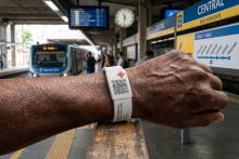 Braço de um homem mostrando pulseira branca de silicone com QR Code. Ao fundo existe uma estação de metrô de Belo Horizonte. Imagem criada com auxílio de Inteligência Artificial